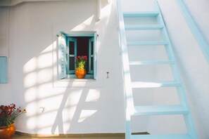Eine weiße Wand mit einem Fenster, ausgestattet mit blauen Fensterläden, einem Topf mit roten Blüten und einer blauen Leiter, die an der Wand lehnt.