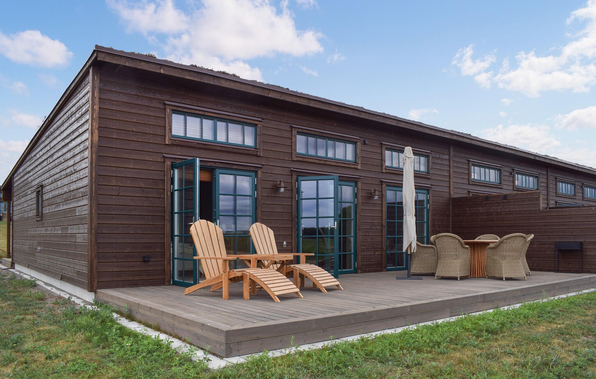A wooden cabin with a deck, two Adirondack chairs, a table, and wicker furniture.