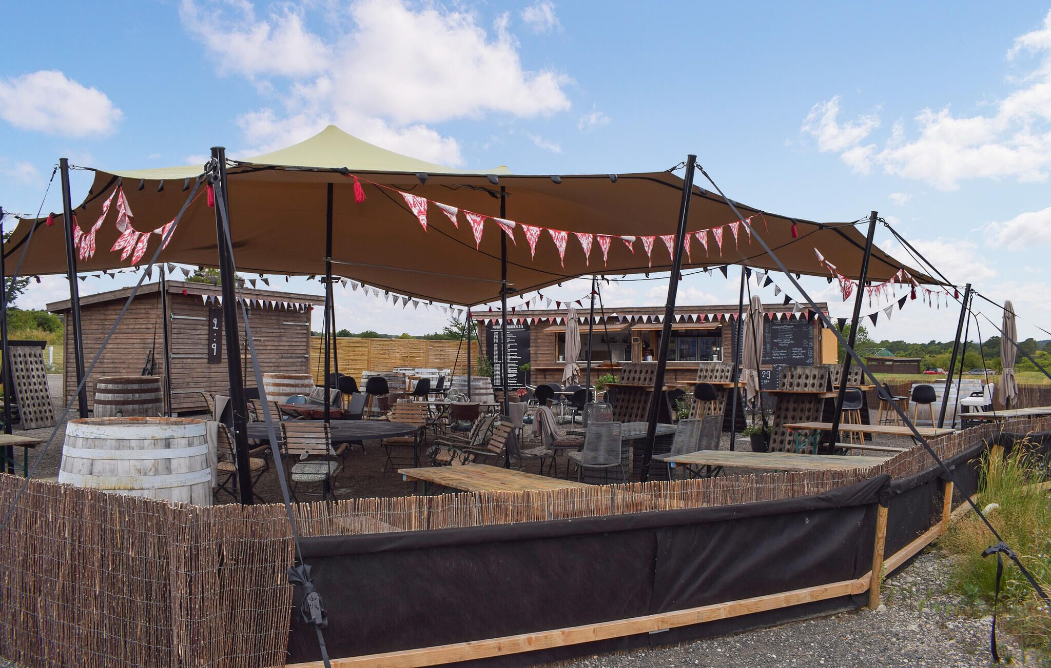 An outdoor seating area with tables and chairs under a large canopy, surrounded by wooden structures and barrels.