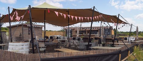 An outdoor seating area with tables and chairs under a large canopy, surrounded by wooden structures and barrels.