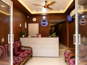 A reception area with a wooden reception desk, a clock, and a sign that reads 'KHGH'. There are two sofas with patterned upholstery and a ceiling fan.