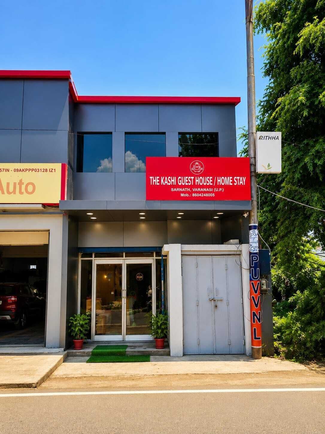 Um edifício com uma placa vermelha indicando 'The Kashi Guest House / Home Stay' e uma porta de garagem visível.