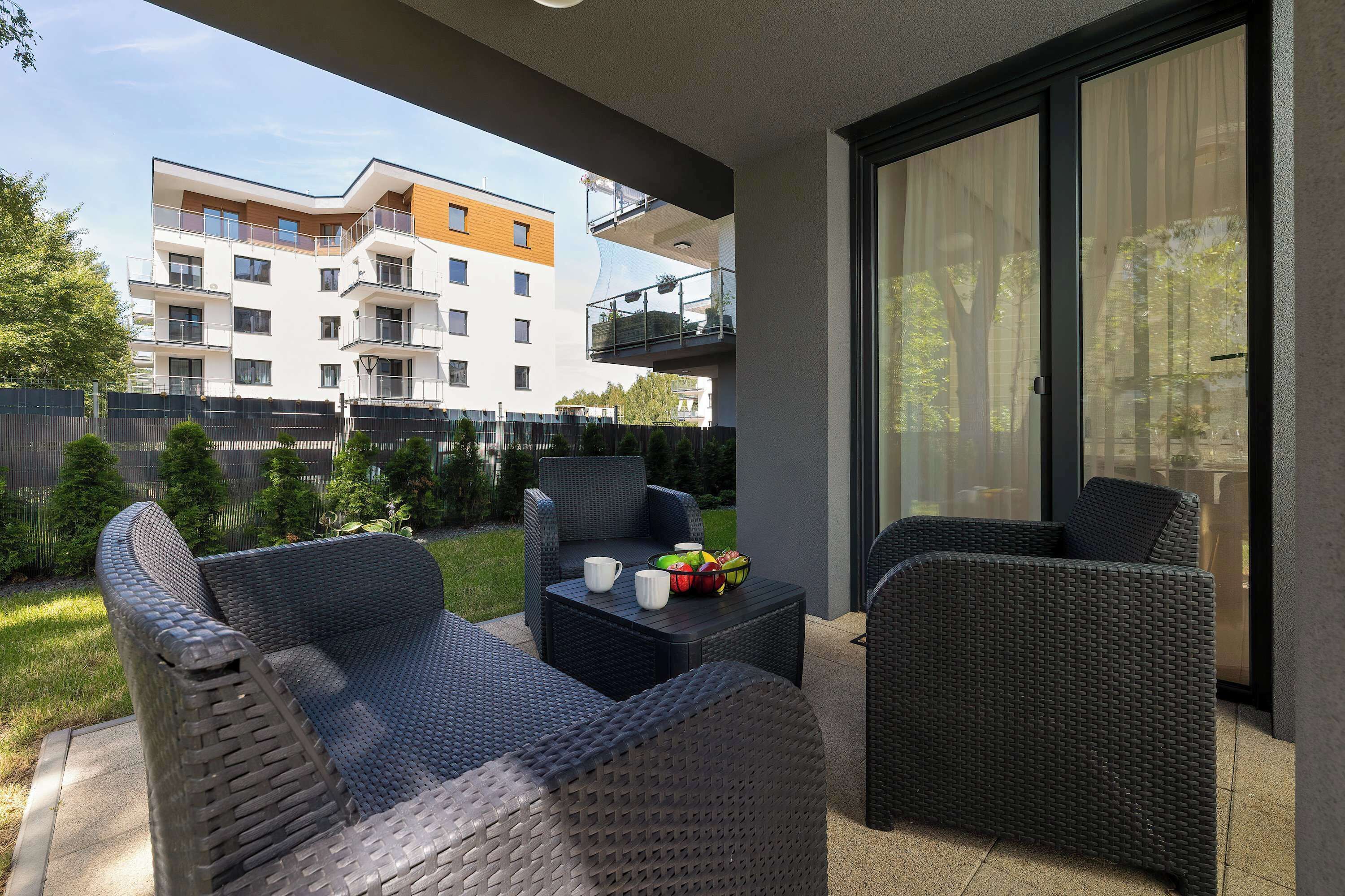 Un espace de détente extérieur moderne, avec des meubles en osier, une table garnie de fruits et de tasses, et une vue sur un immeuble à plusieurs étages en arrière-plan.