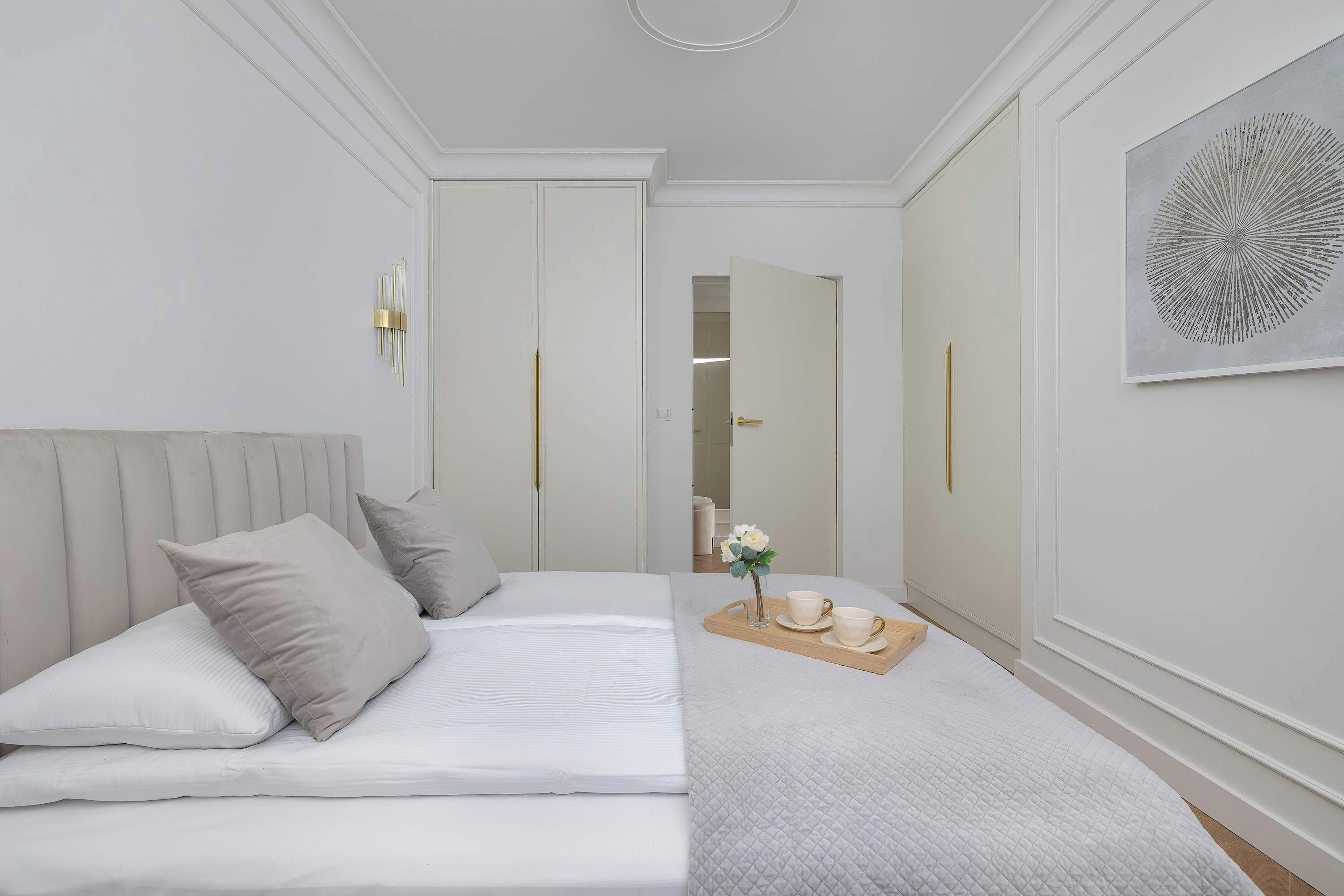Quarto de hotel com cama, mesa de cabeceira com um vaso e chávenas, e um quadro abstrato grande na parede.