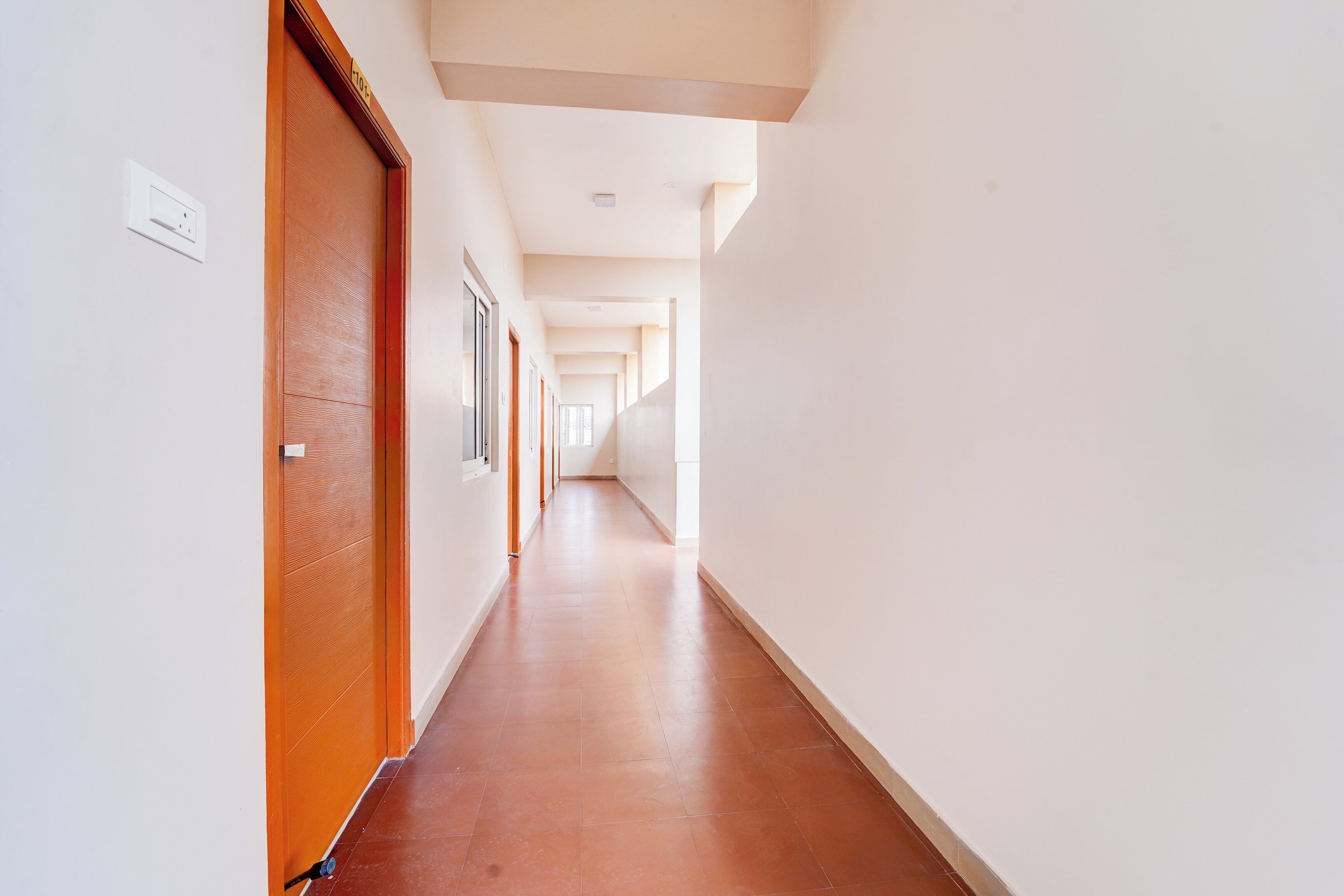 Un largo pasillo estrecho con puertas de madera a un lado y ventanas al otro, que conduce a una habitación luminosa al final.