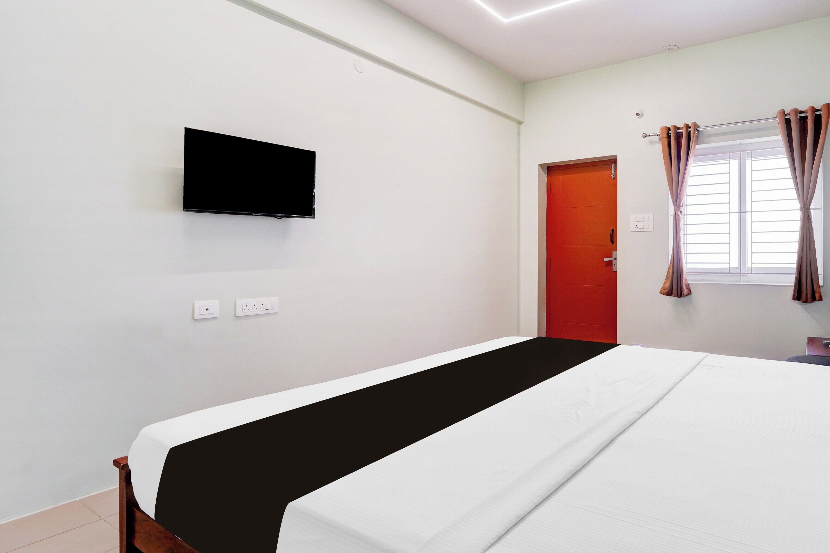 A hotel room with a bed, a television mounted on the wall, a red door, and a window with curtains.