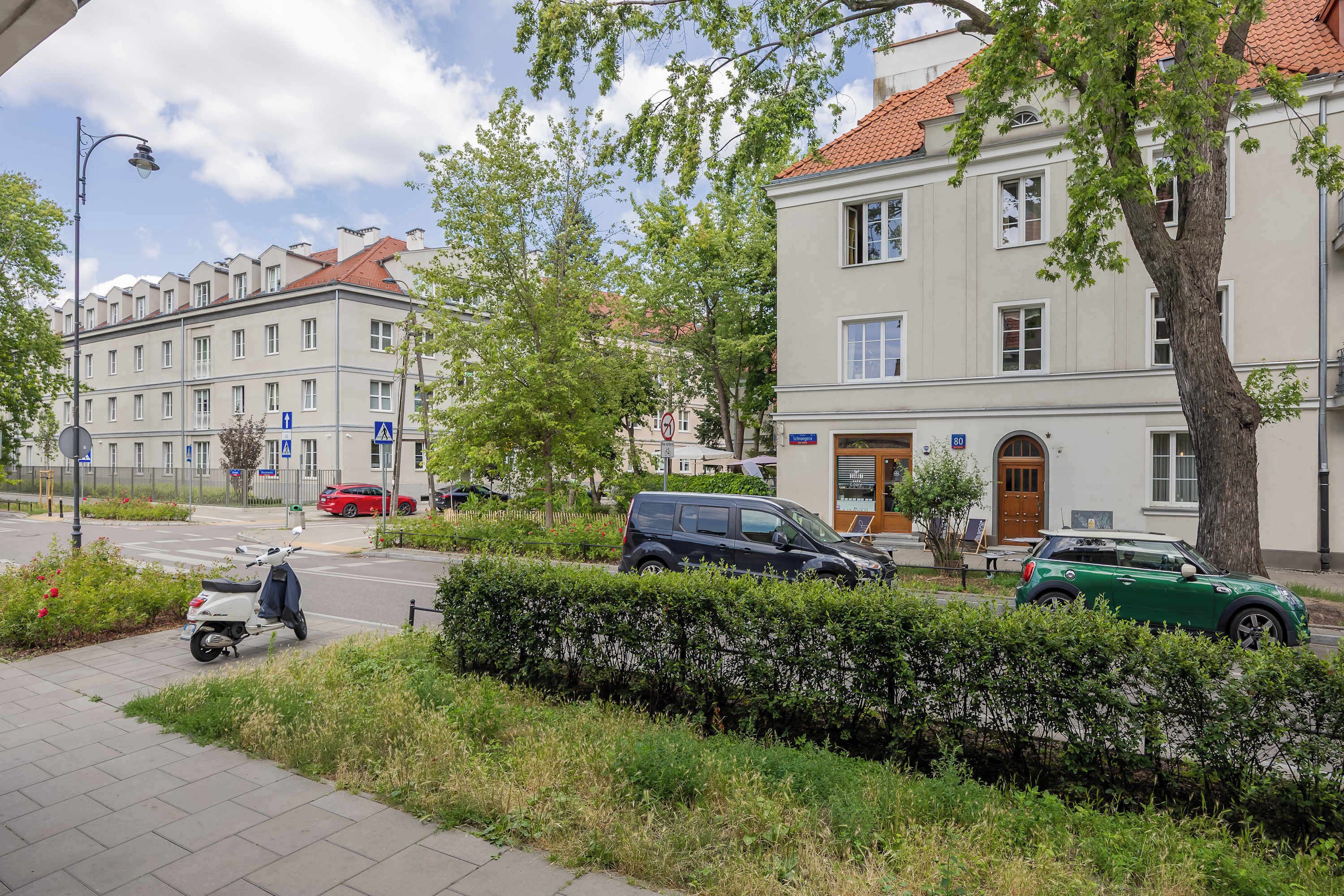 Et gadesyn med parkerede biler, en knallert og en bygning med et skilt der står 'Schroeger'.