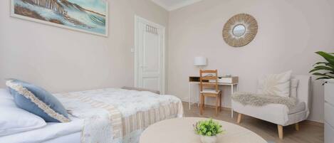 Un dormitorio con una cama, una silla, un escritorio, una mesa redonda con una planta y un cuadro en la pared.