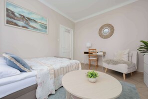 A bedroom with a bed, a chair, a desk, a round table with a plant, and a painting on the wall.