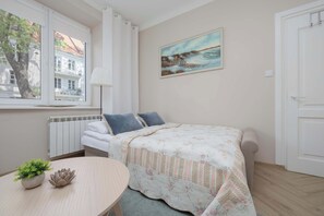 A bedroom with a bed, a painting, a radiator, a window, and a table with a plant. - Cozy Studio with a Workspace | Near the Metro (Warszawa)