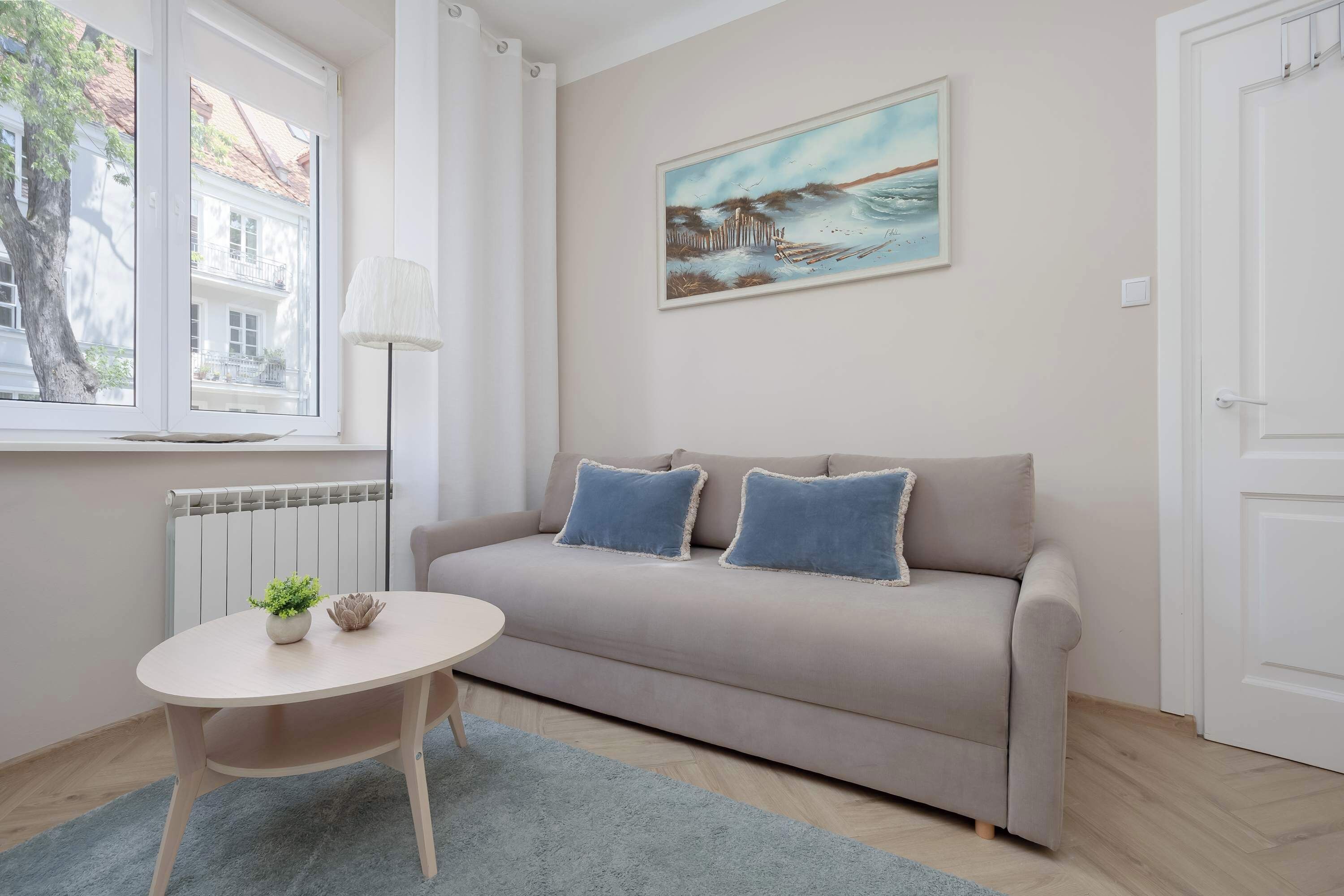 Ein modernes Wohnzimmer mit einem beigen Sofa, zwei blauen Kissen, einem kleinen runden Tisch mit Pflanze und Dekoration, einer Stehlampe und einem Gemälde mit Küstenmotiv.