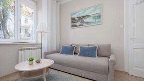Ein modernes Wohnzimmer mit einem beigen Sofa, zwei blauen Kissen, einem kleinen runden Tisch mit Pflanze und Dekoration, einer Stehlampe und einem Gemälde mit Küstenmotiv.