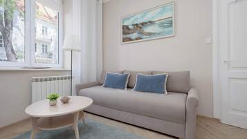 Ein modernes Wohnzimmer mit einem beigen Sofa, zwei blauen Kissen, einem kleinen runden Tisch mit Pflanze und Dekoration, einer Stehlampe und einem Gemälde mit Küstenmotiv.