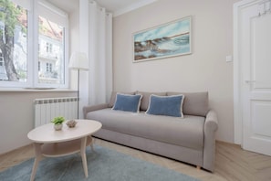 A modern living room with a beige sofa, two blue pillows, a small round table with a plant and a decorative item, a floor lamp, and a painting of a coastal scene. - Cozy Studio with a Workspace | Near the Metro (Warszawa)