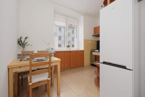 A compact kitchen with a white refrigerator, wooden cabinets, a window overlooking a building, and a dining table with chairs. - Cozy Studio with a Workspace | Near the Metro (Warszawa)