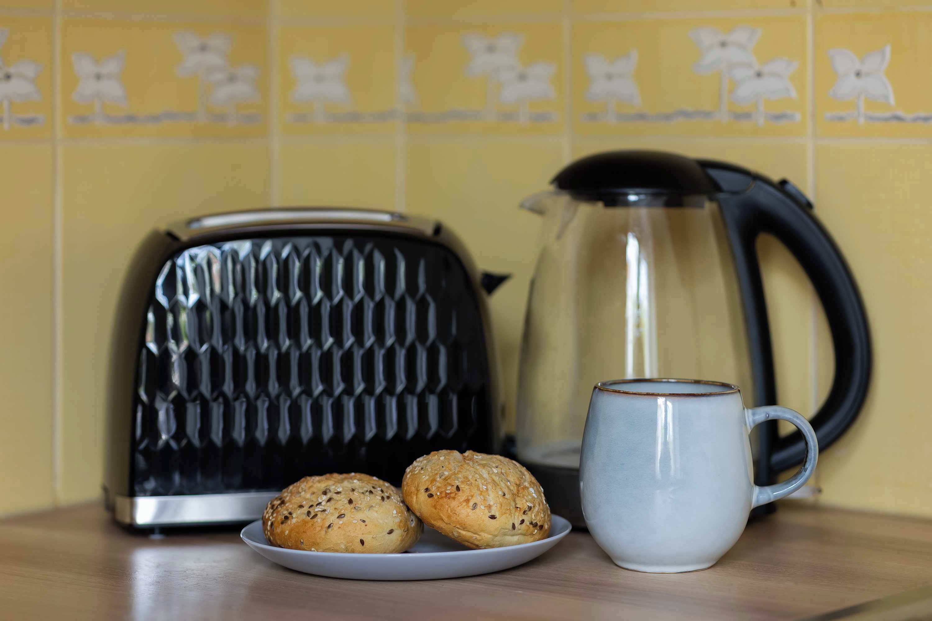 A kitchen counter with a toaster, a kettle, a plate with two rolls, and a mug.