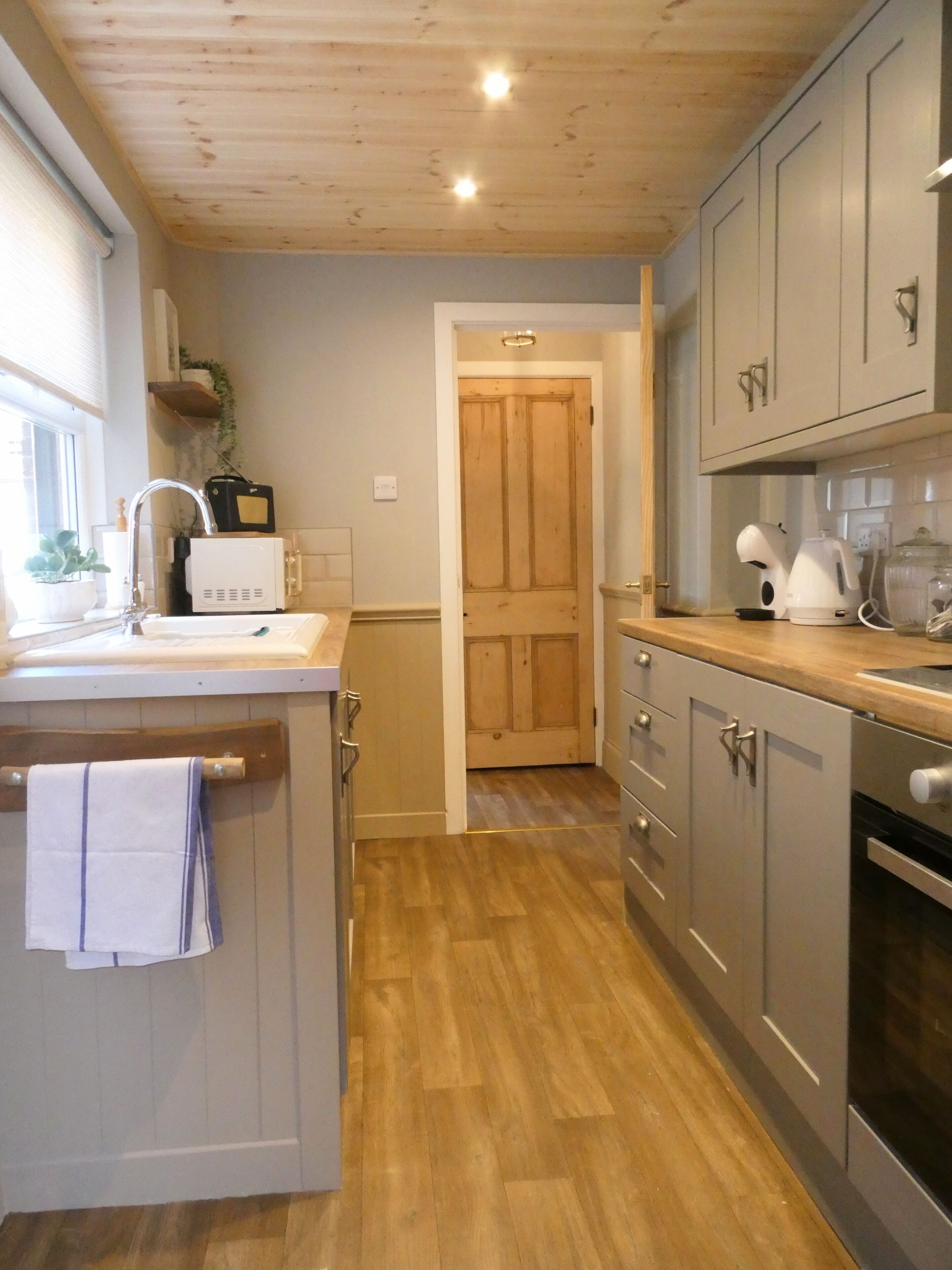 A kitchen with wooden flooring, a sink, cabinets, and a door leading to another room.