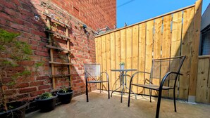 Un espacio patio con pared de ladrillo, cerca de madera, sillas de metal y plantas en macetas.
