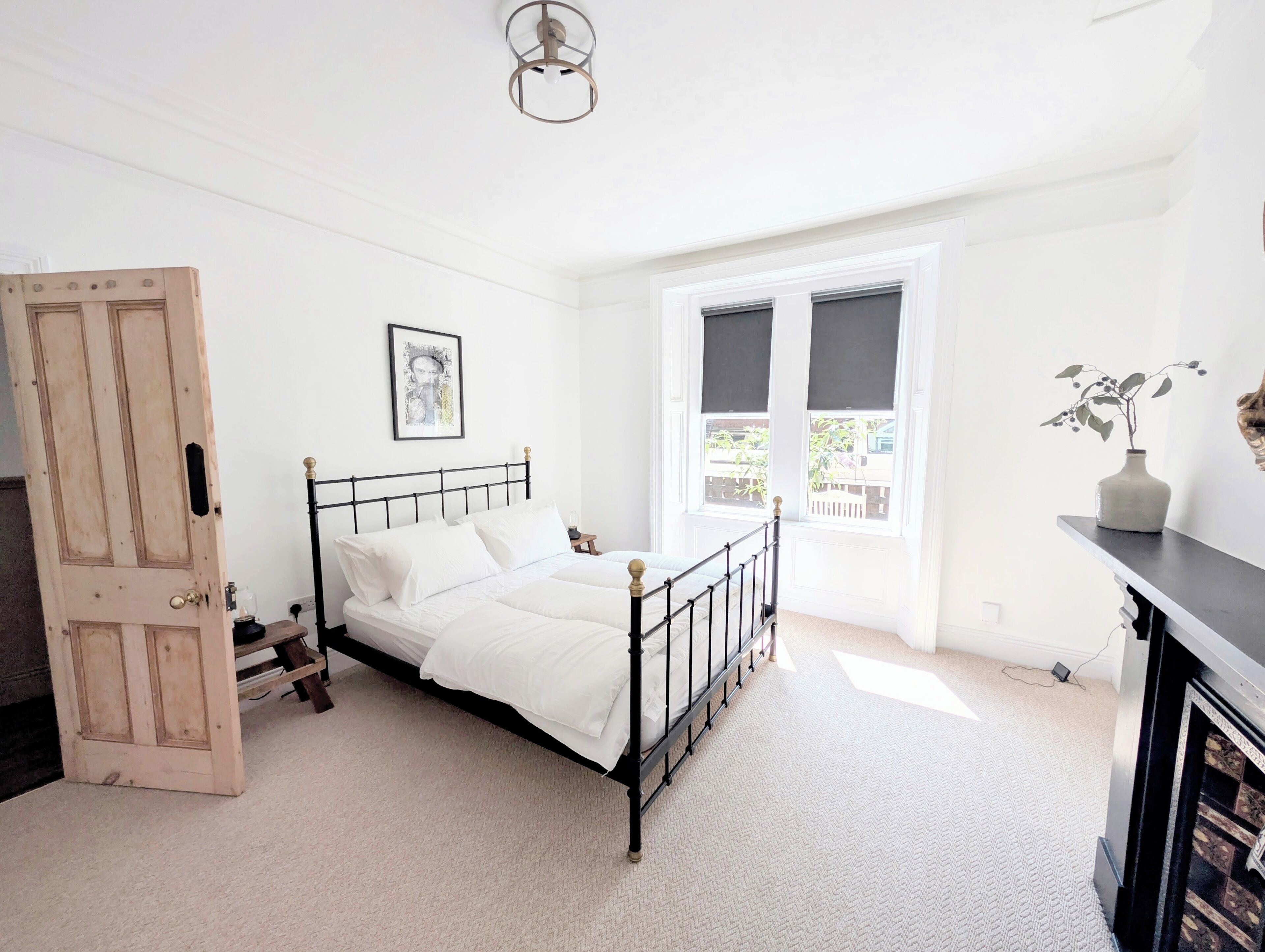 A bedroom with a bed, a door, a window, a picture on the wall, and a vase with plants.