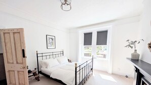 Un dormitorio con cama, una puerta, una ventana, un cuadro en la pared y un florero con plantas.