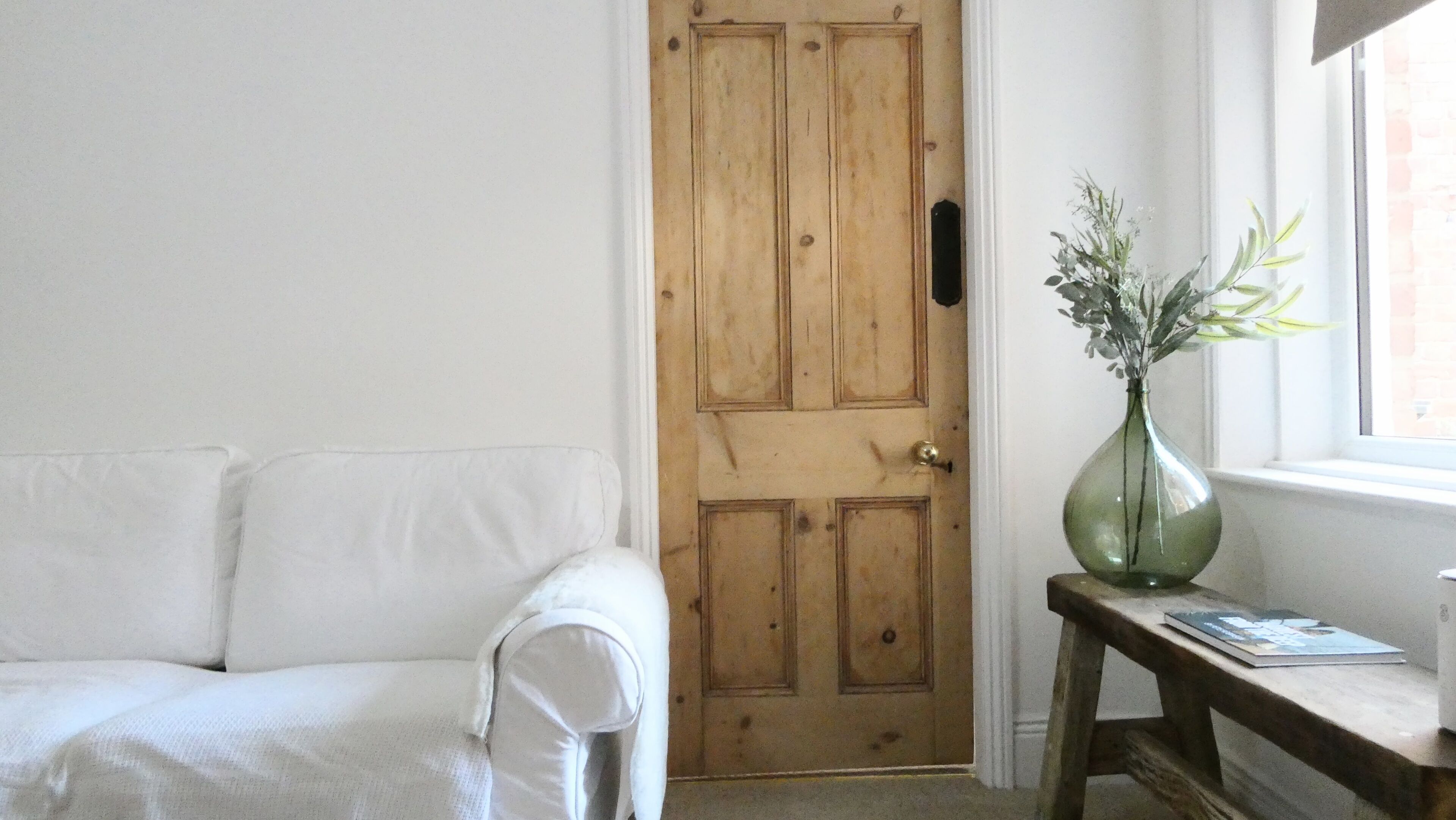 A room with a wooden door, a white sofa, and a small table with a vase.