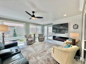 A spacious living room with a fireplace, ceiling fan, and large windows. - Coral Crest at Frogmore - 3/3 w/ 4-Seat Golf Cart in The Villages - Moultrie Creek (Moultrie Creek, The Villages)