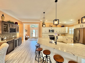 A modern kitchen with a central island, bar stools, and a dining area with a sofa. - Coral Crest at Frogmore - 3/3 w/ 4-Seat Golf Cart in The Villages - Moultrie Creek (Moultrie Creek, The Villages)