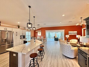 A modern kitchen with a central island, bar stools, and a dining area with a sofa and chairs. - Coral Crest at Frogmore - 3/3 w/ 4-Seat Golf Cart in The Villages - Moultrie Creek (Moultrie Creek, The Villages)