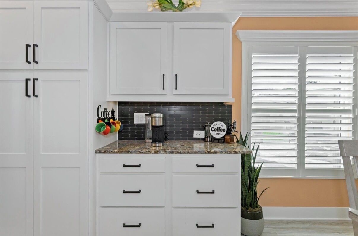 A kitchen with white cabinets, a coffee station, and a window with blinds.