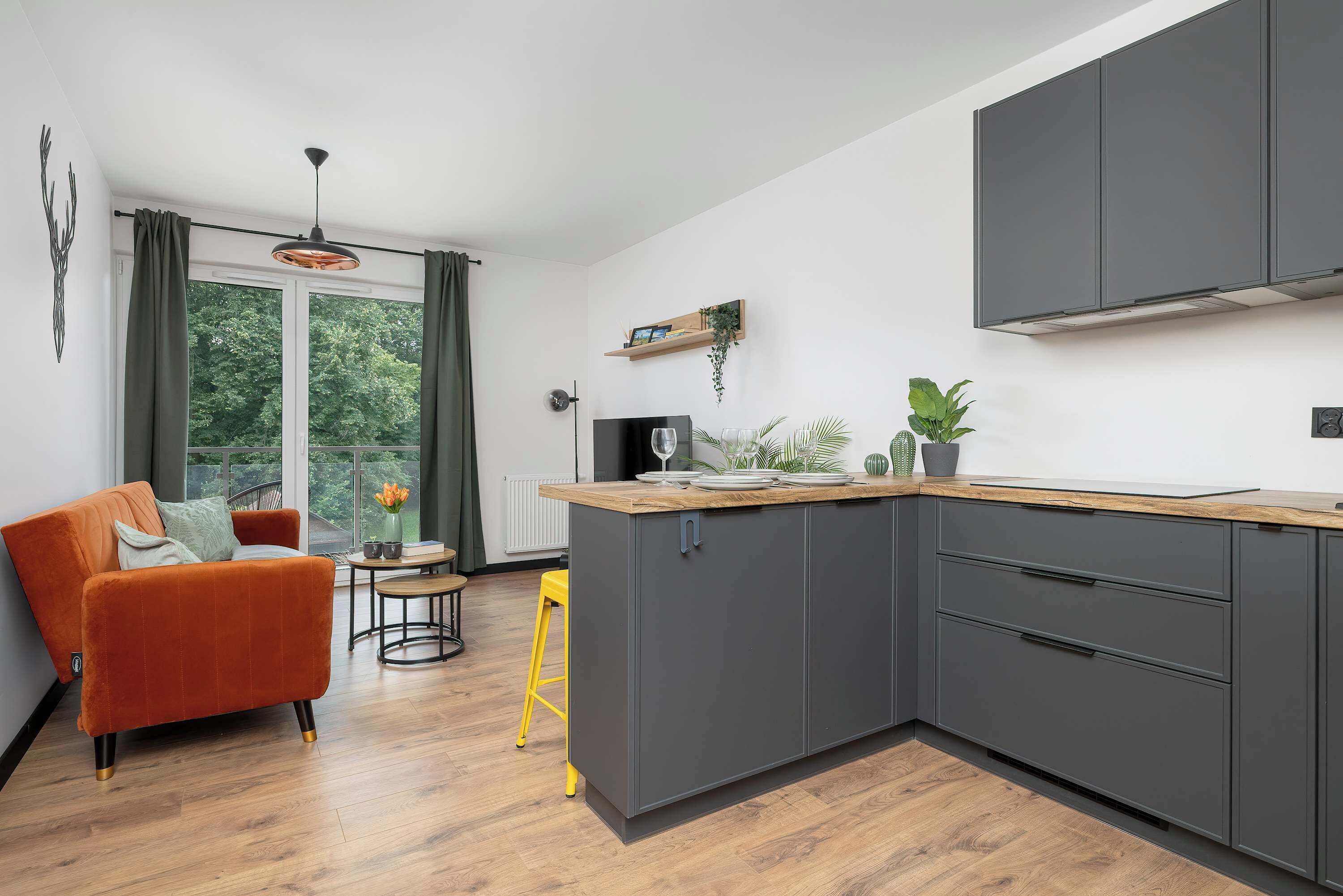 Une cuisine moderne avec des meubles gris, un plan de travail en bois et un coin repas agrémenté d’un canapé rouge et d’un tabouret jaune.