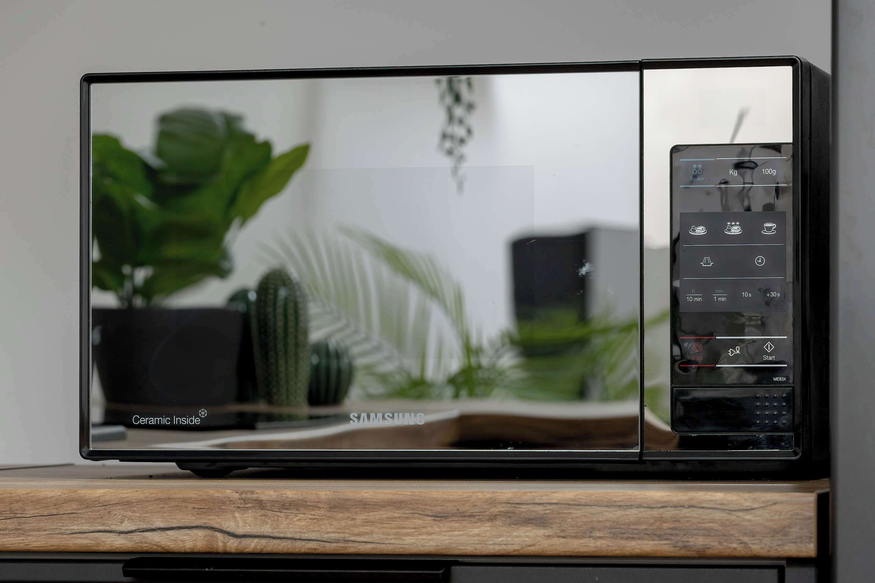 A Samsung microwave on a wooden surface with a reflection of a plant and a white wall.