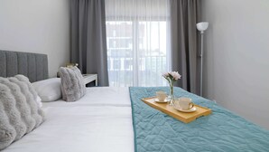 Et hotelværelse med seng, sengebord, gardiner, et vindue med udsigt samt et bakke med kopper og blomster.