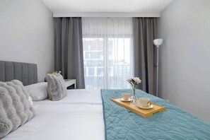 Ein Hotelzimmer mit Bett, Nachttisch, Vorhängen, einem Fenster mit Blick und einem Tablett mit Tassen und Blumen.