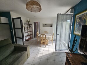 A living room with a sofa, a dining table with chairs, a bookshelf, and a large window with a curtain. - L'amirauté pour 4 personnes (La Rochelle)