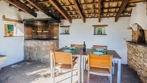 An outdoor dining area with a stone wall, wooden ceiling beams, and a built-in grill. - Hillside Villa With Pool for 5 (Pollença)