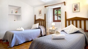 A room with two single beds, a small bedside table with a lamp, a window with a view of greenery, and a shelf with decorative items. - Hillside Villa With Pool for 5 (Pollença)
