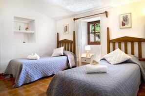 A room with two single beds, a small bedside table with a lamp, a window with a view of greenery, and a shelf with decorative items. - Hillside Villa With Pool for 5 (Pollença)