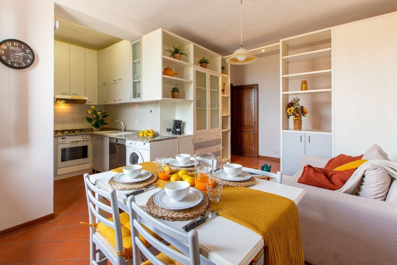 A compact kitchen with white cabinets, a dining table set for two, and a sofa in the adjacent living area.