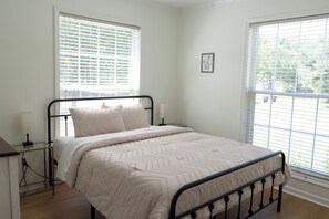 A bedroom with a bed, a bedside table, a lamp, and two windows with blinds.