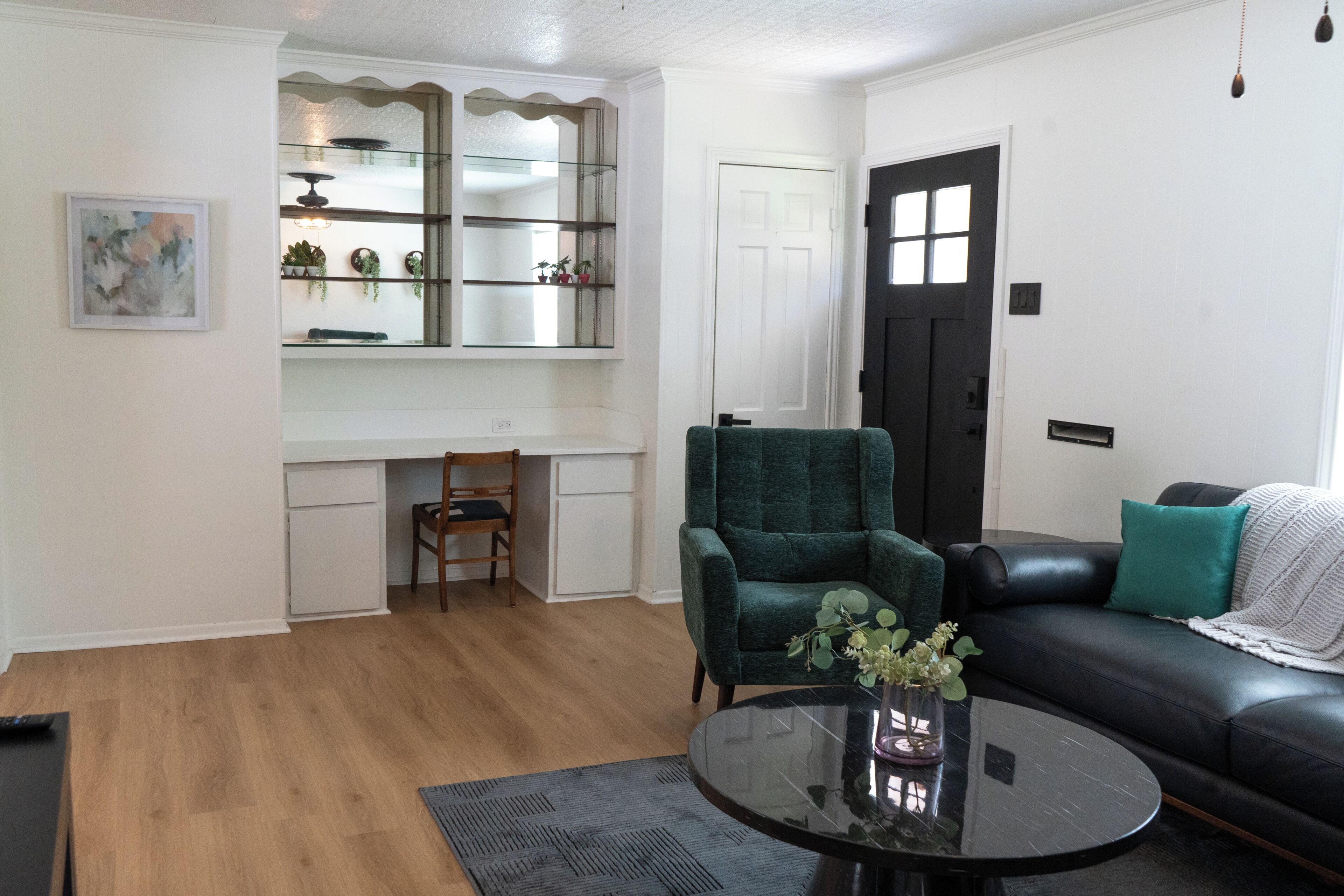 Un salon comprenant un bureau, un fauteuil vert, un canapé noir et une petite table ronde sur laquelle est posé un vase.