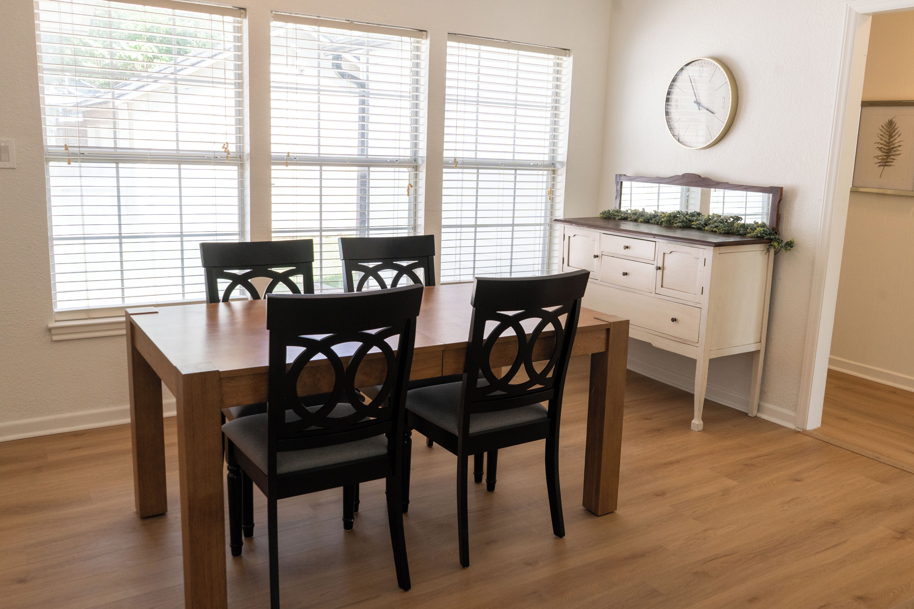 Un coin repas avec une table en bois et quatre chaises, un meuble blanc avec une horloge, et une fenêtre avec des stores.