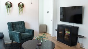 A living room with a teal armchair, a glass coffee table with a vase, a flat-screen TV on a cabinet, and a small side table. - This home located in the Temple Historic District has recently been renovated and updated for a luxurious stay, while maintaining the aesthetic historical  vibe. (TEMPLE)