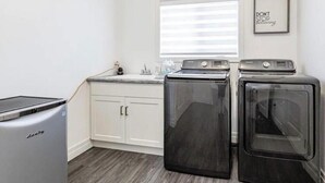A laundry room with two front-loading washers and a dryer, a small countertop with a sink, and a window with blinds.