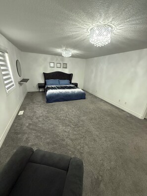 A bedroom with a bed, a small bedside table, a nightstand, a chandelier, and a window with blinds.