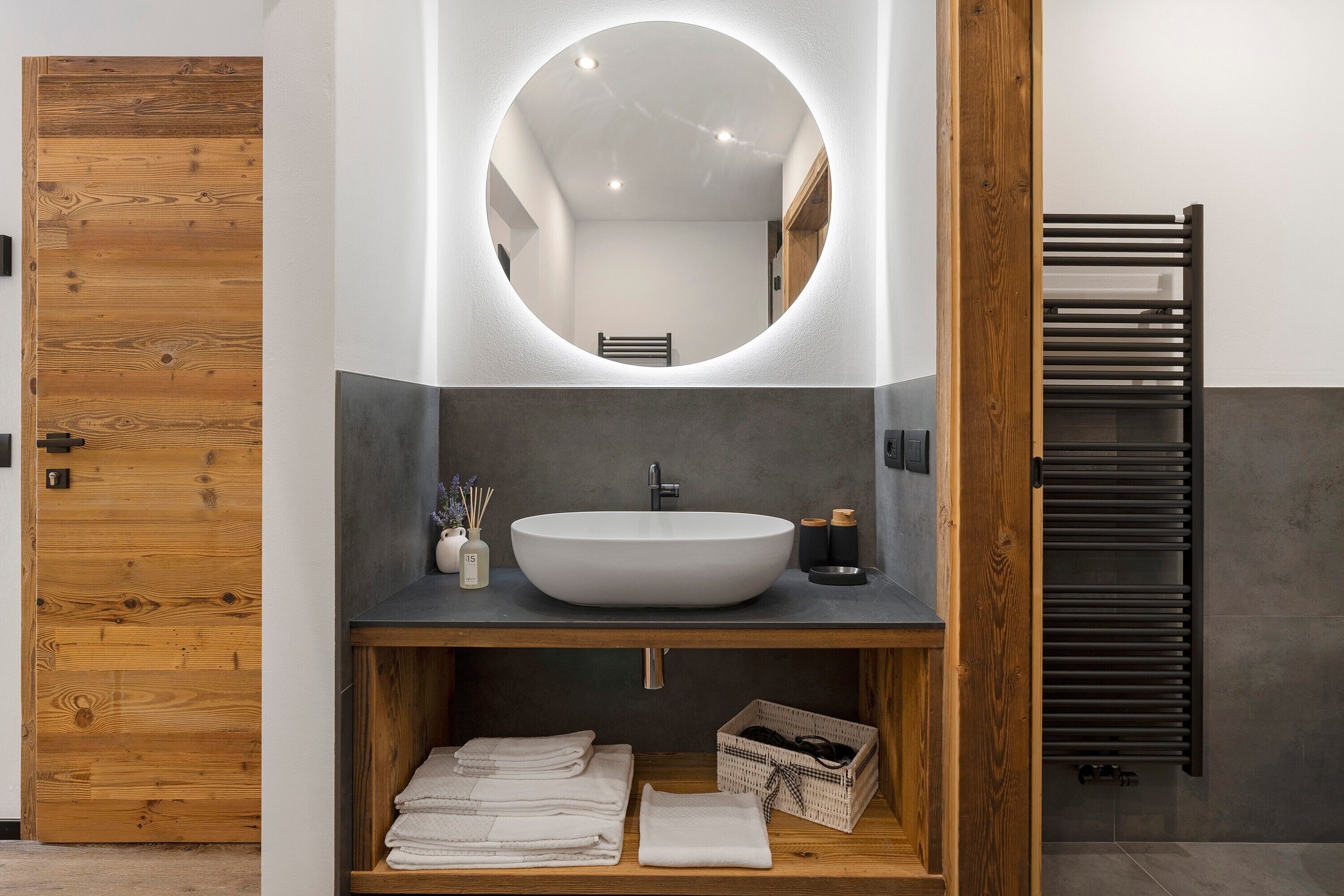 Un baño moderno con puerta de madera, espejo redondo, lavamanos tipo recipiente y espacio para toallas.