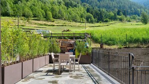 Een terras buiten met stenen vloer, een tafel en stoelen, en uitzicht op een groen landschap met bomen en een heuvel op de achtergrond.
