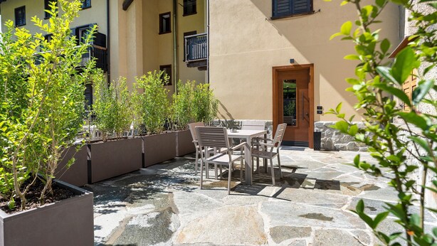 En stenbelagt terrasse med udendørs møbler, omgivet af plantekasser og en bygning med flere vinduer.