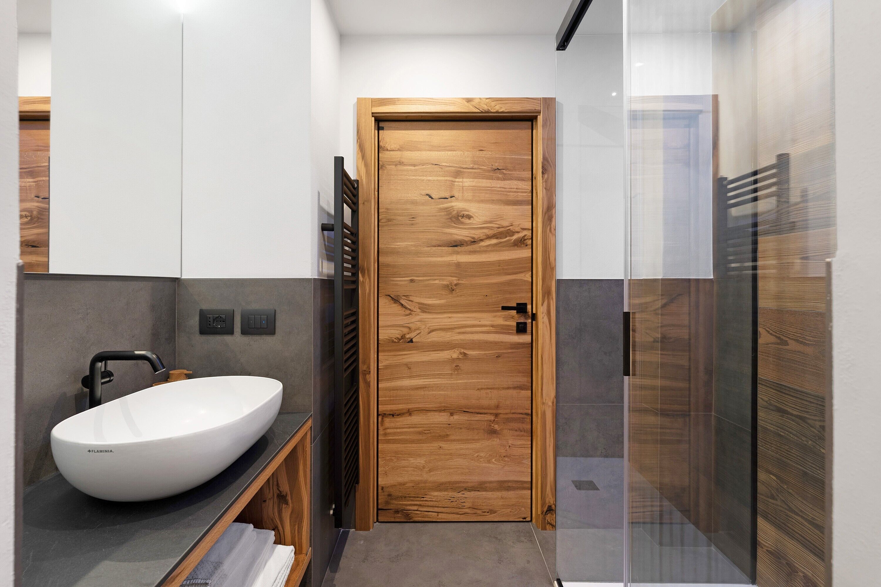 Un bagno moderno con una porta in legno, un lavabo a incasso bianco, un portasciuga e una cabina doccia in vetro.