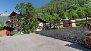 Un edificio con pared de piedra y valla de madera, camino de entrada pavimentado y montañas al fondo.