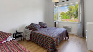Ein Schlafzimmer mit zwei Betten, einem Fenster mit Blick auf Grünflächen, einem Nachttisch mit Lampe und einer Heizung.